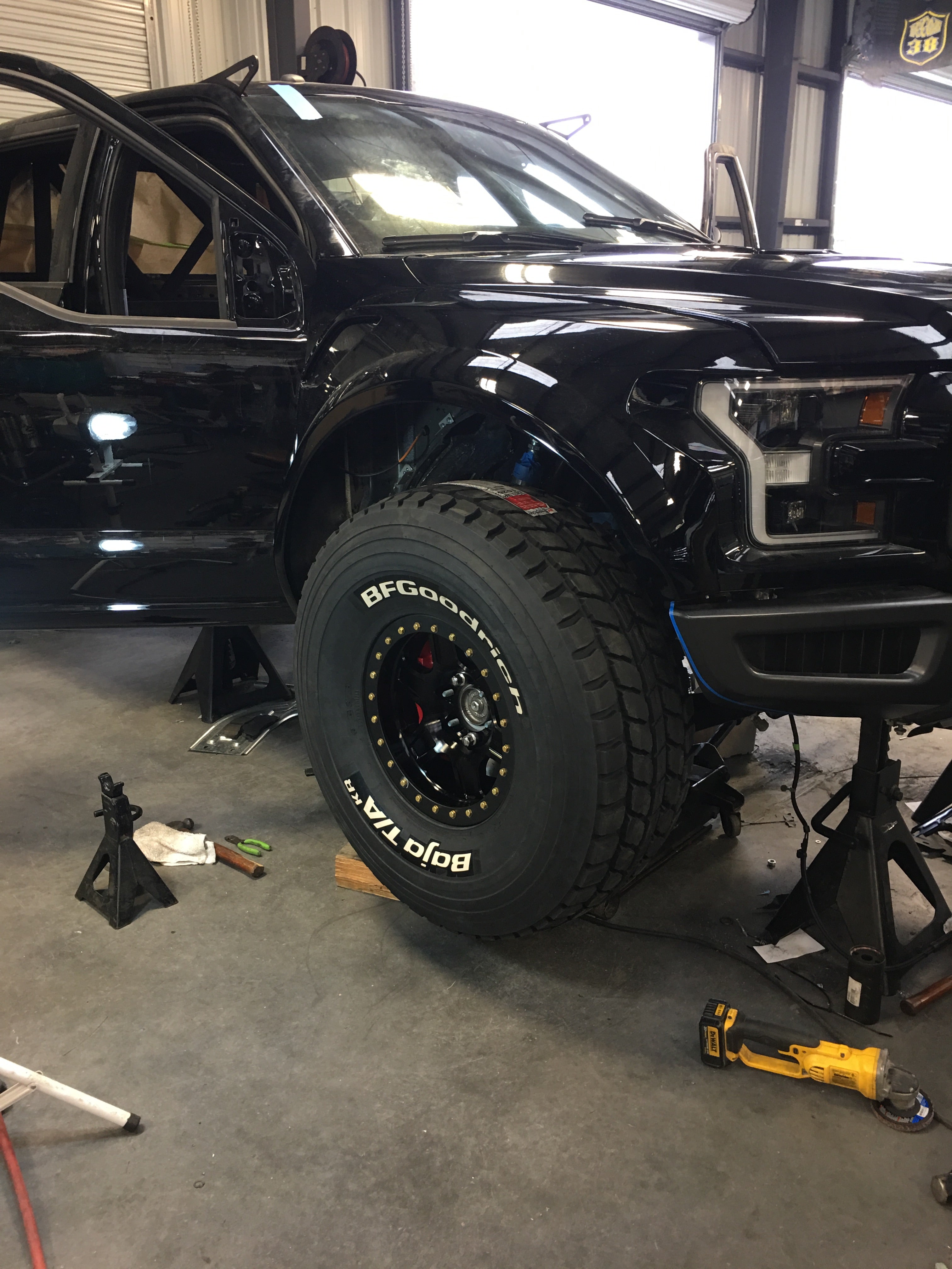 2017 Up Raptor Long Travel Front Suspension Kit installed on a black Ford Raptor with large off-road tires in a workshop.