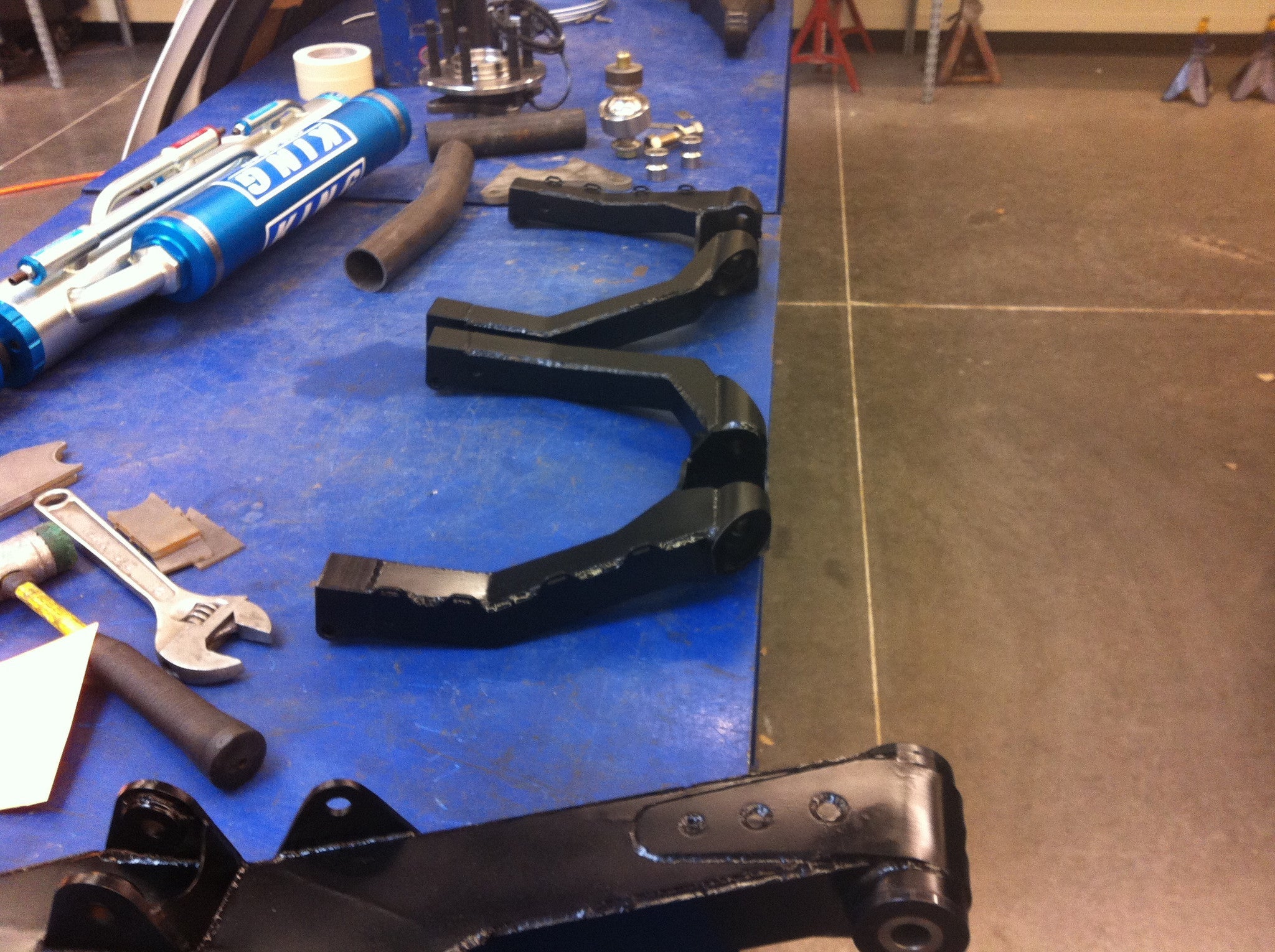 Long travel front suspension kit components for 2010-2014 Raptor, featuring black metal arms and various tools on a workbench.