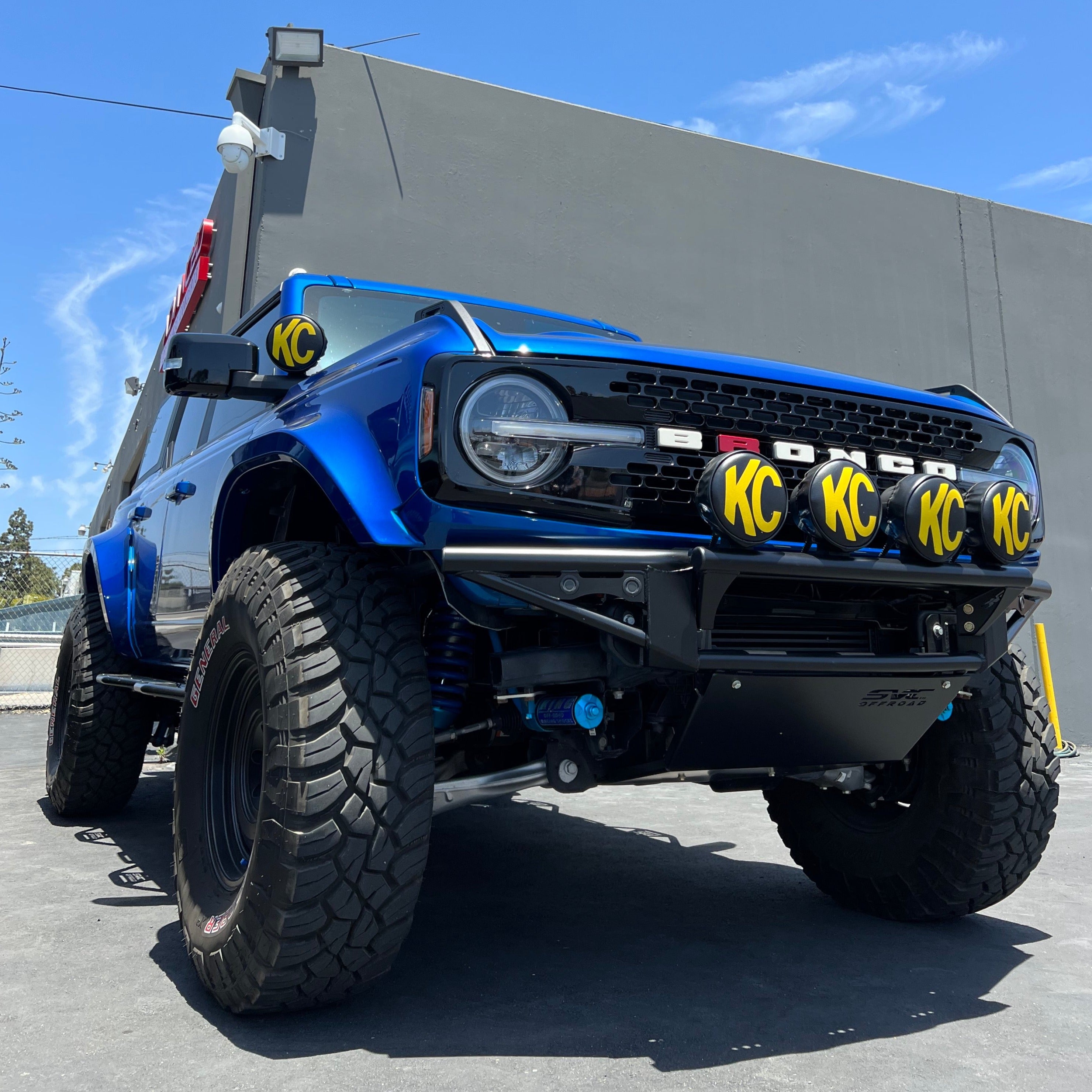 2021-2022 Ford Bronco 4-Door with a rear quarter kit, featuring off-road tires and KC lights, parked against a gray wall.
