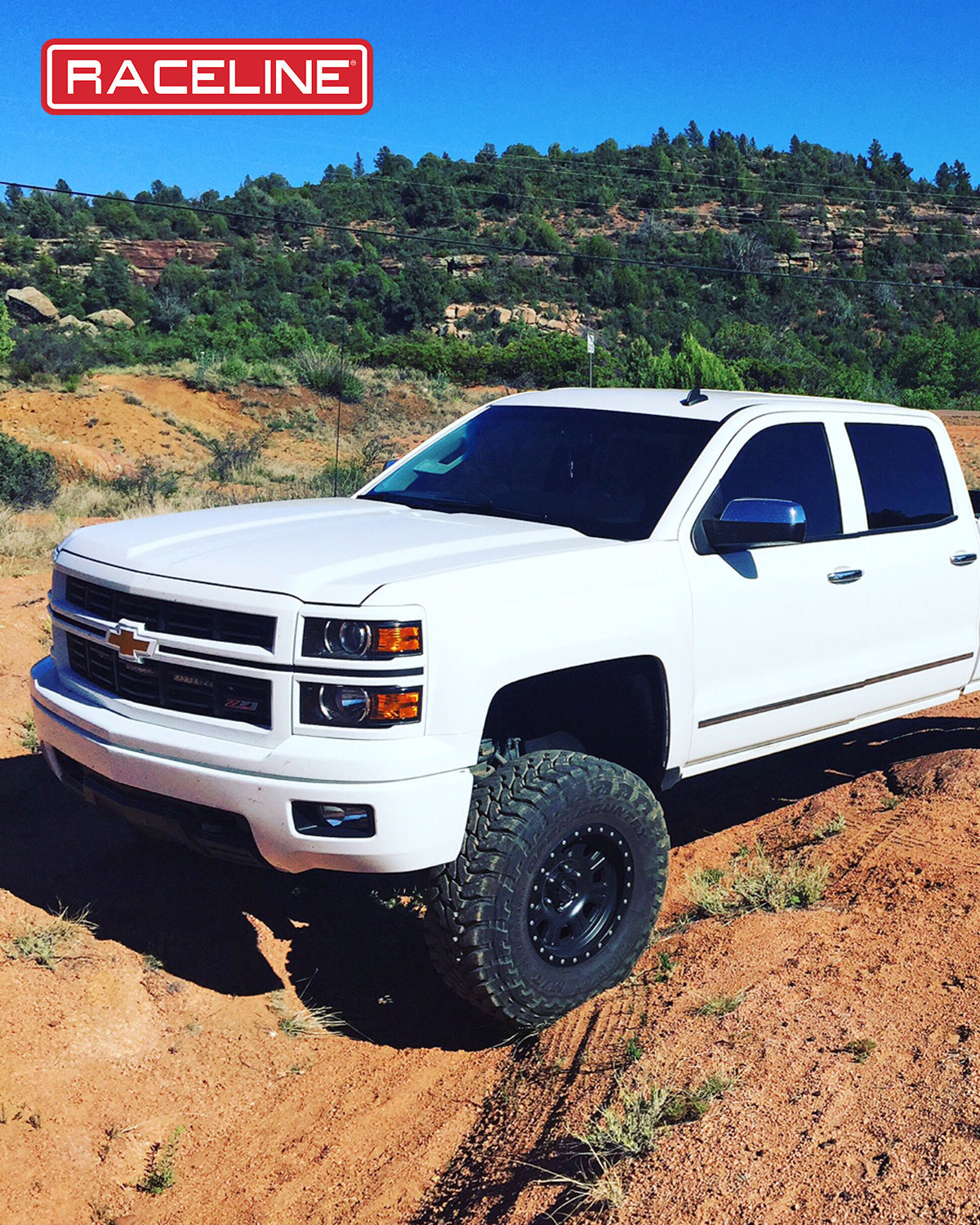 White Chevrolet truck equipped with Raceline 928B Monster wheels, parked on a dirt trail surrounded by greenery.