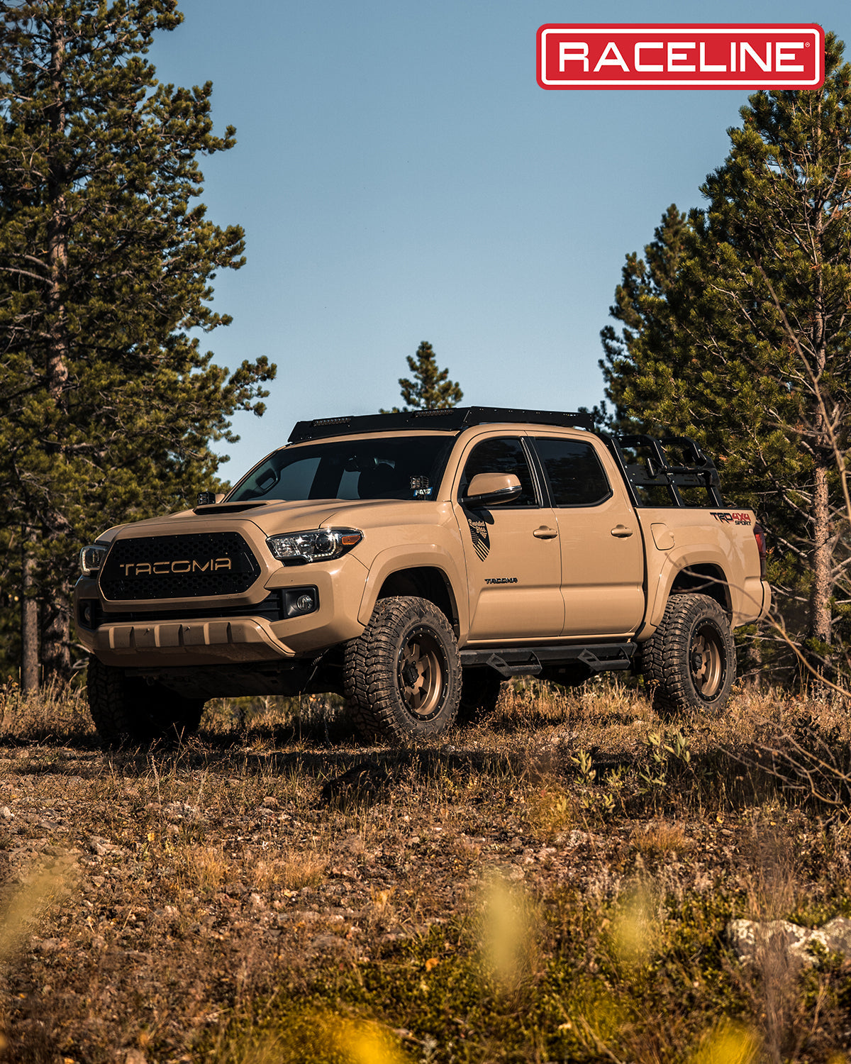 Tan Tacoma truck equipped with 935BZ Defender wheels, parked in a forested area with tall trees.