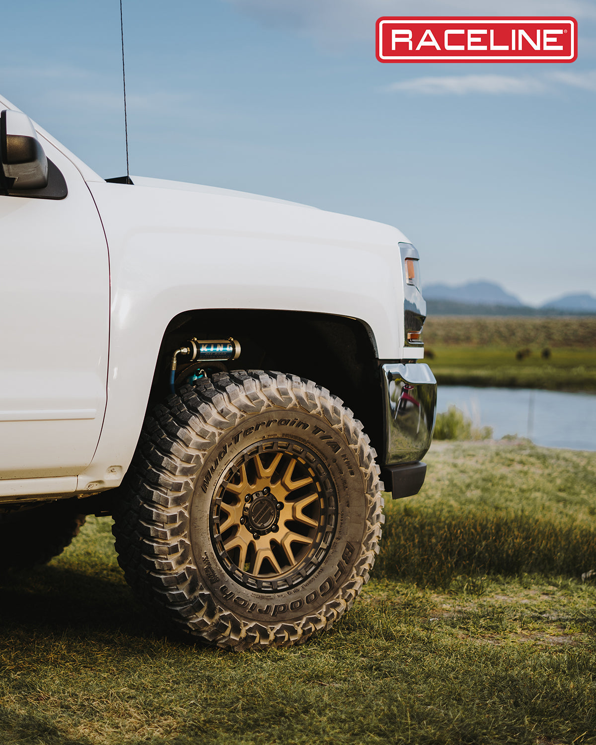 Close-up of a white truck's wheel featuring a Raceline 953BZ Krank rim and rugged tire, set against a scenic outdoor backdrop.
