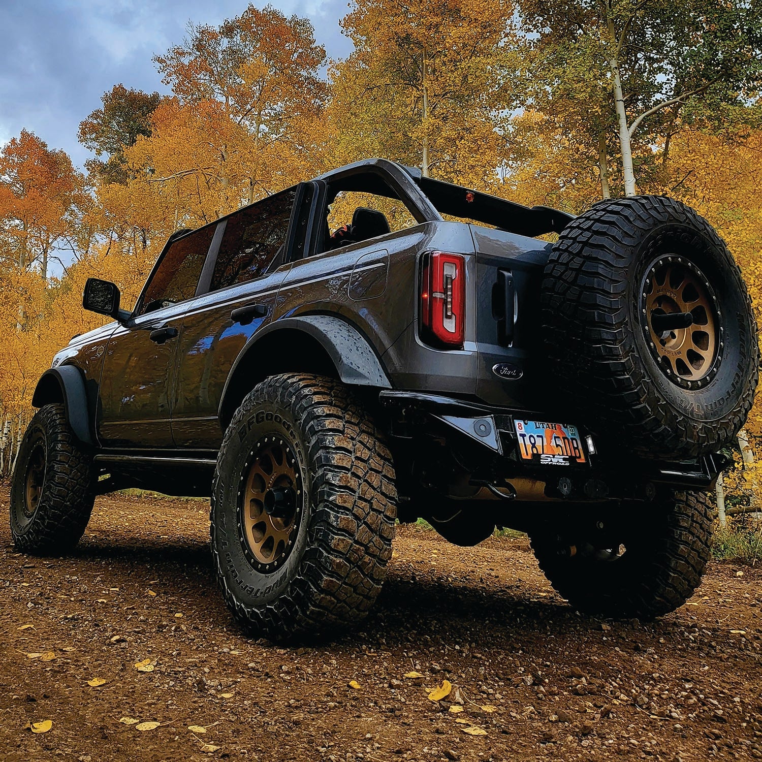2021 Ford Bronco with Baja rear bumper parked on a dirt trail surrounded by autumn foliage.
