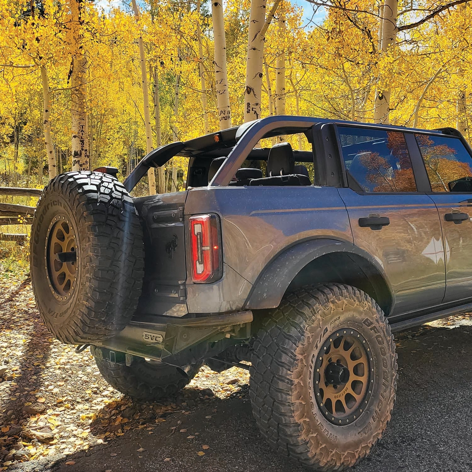 2021 Ford Bronco with Baja rear bumper and oversized tire, parked in a scenic autumn landscape with yellow foliage.