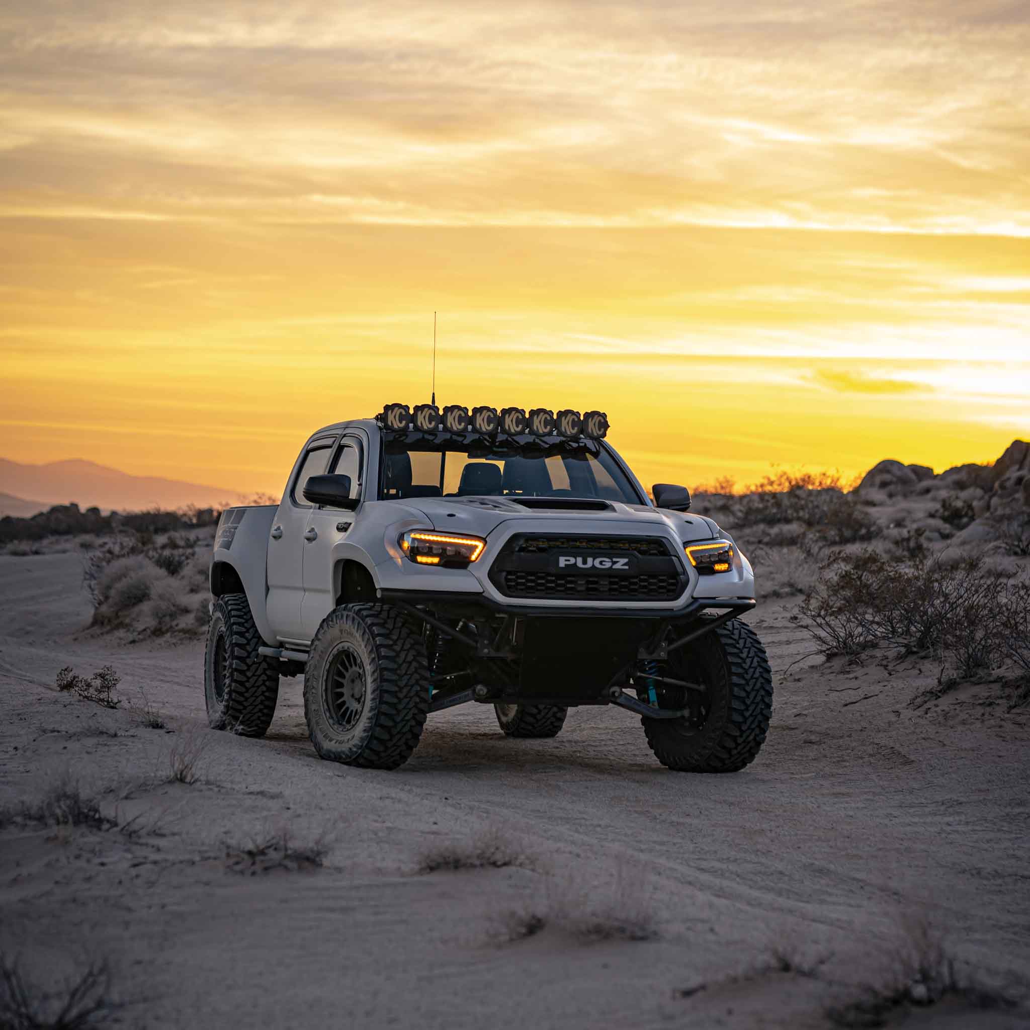 2016-2023 Toyota Tacoma with aftermarket fenders and off-road lights in a desert landscape at sunset.