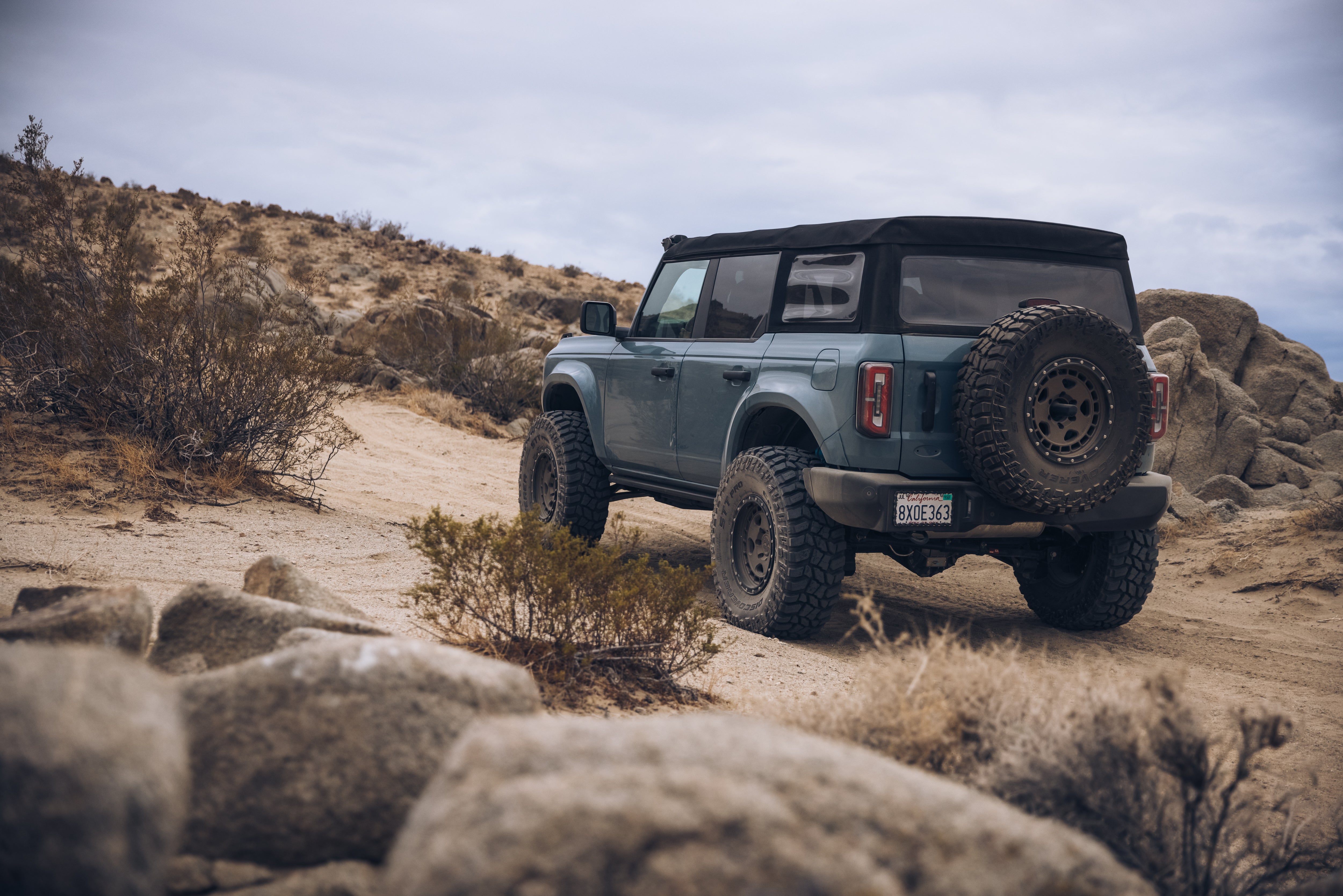 2021 Ford Bronco with ADV fiberglass parts driving on a rocky off-road trail in a desert landscape.