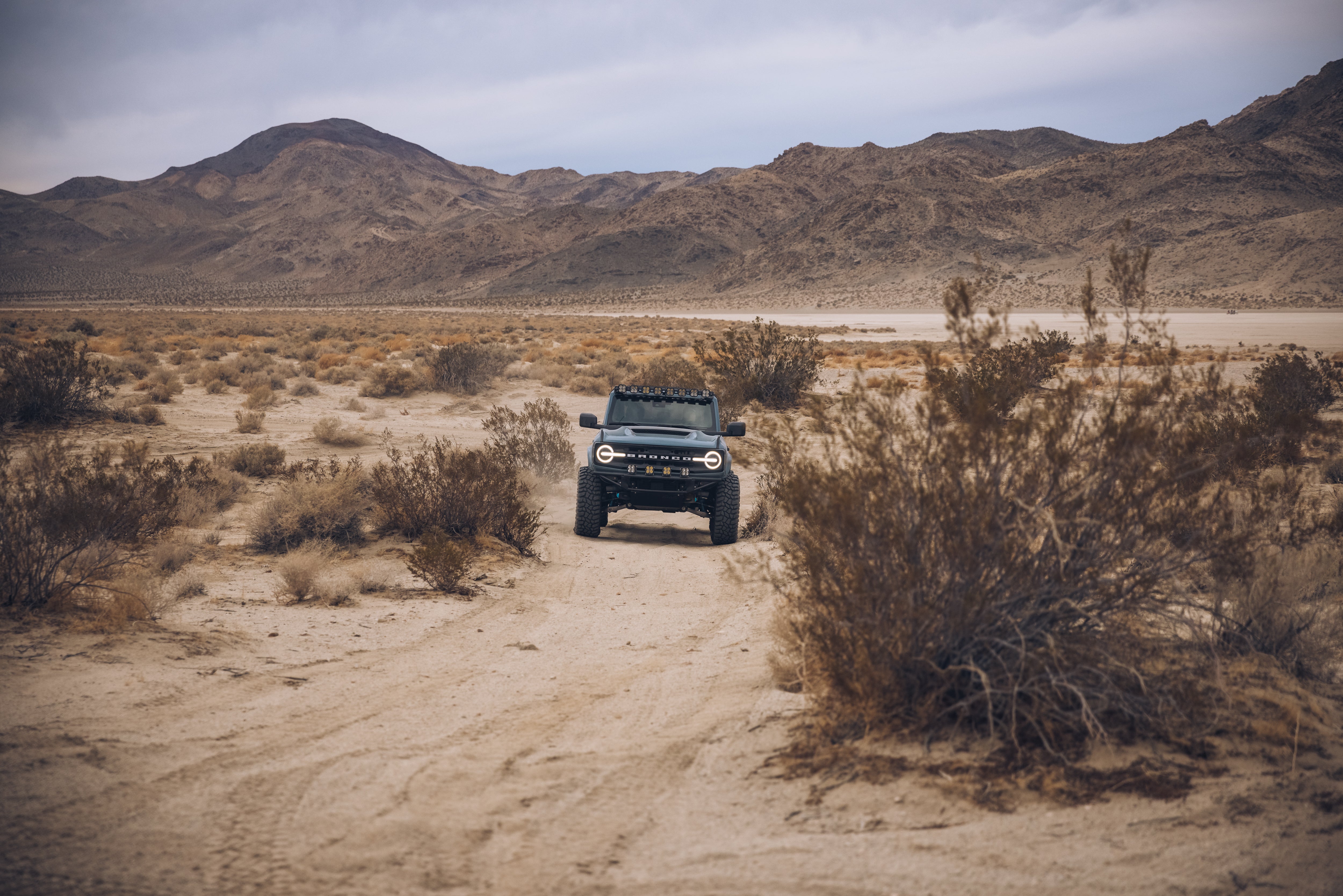 2021 Ford Bronco with ADV fiberglass parts driving on a sandy off-road trail in a desert landscape.