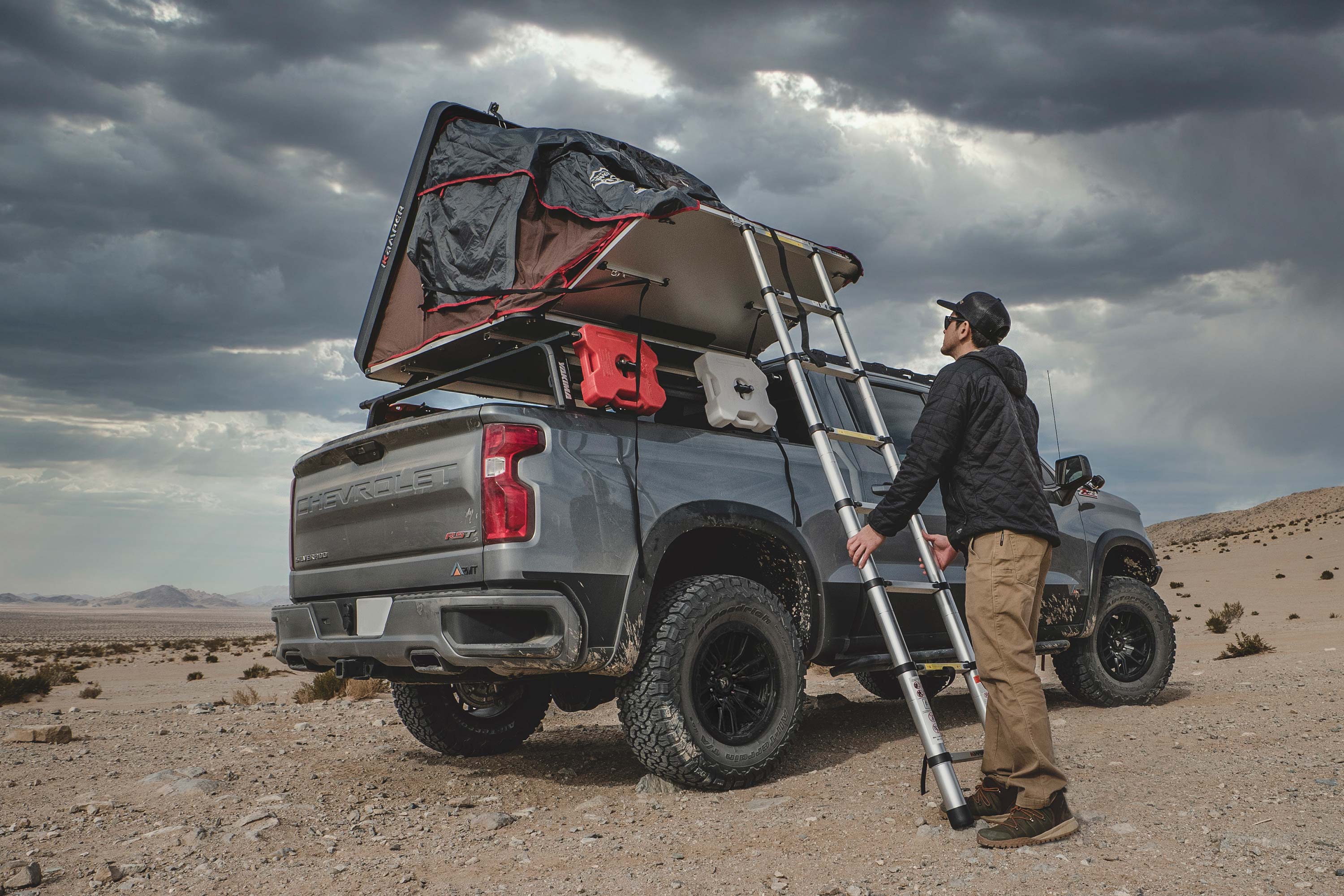 A man sets up a rooftop tent on a lifted Chevy Silverado with a FOX Performance Elite Coil-Over lift kit in a desert landscape.