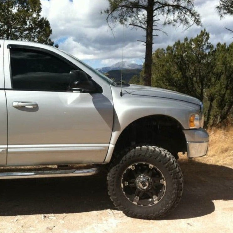 Silver 2003-2005 Dodge Ram with aftermarket fenders and large off-road tires parked in a natural setting.
