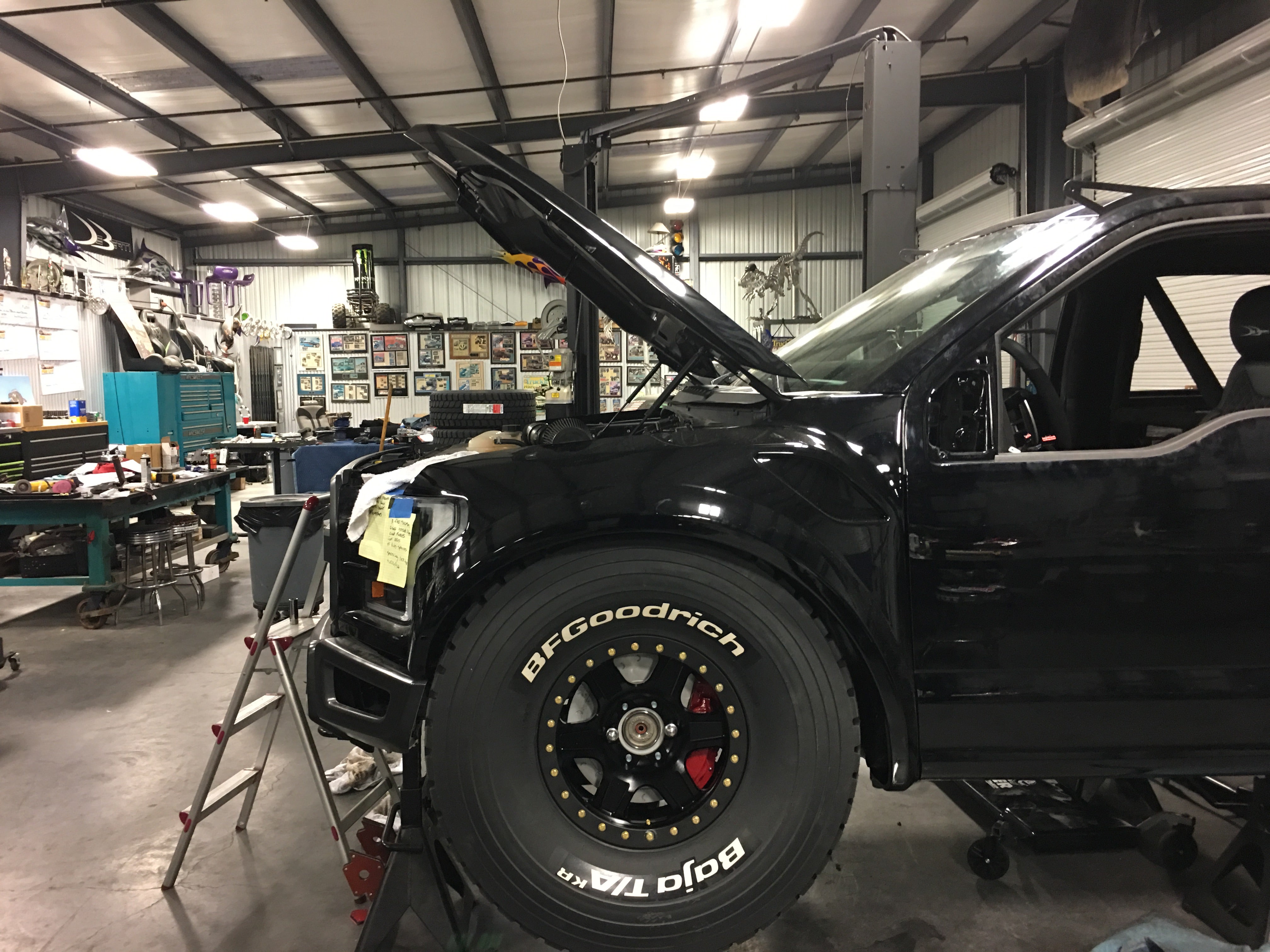 2017 Up Raptor Long Travel Front Suspension Kit installed on a black truck in a workshop with tools and equipment in the background.