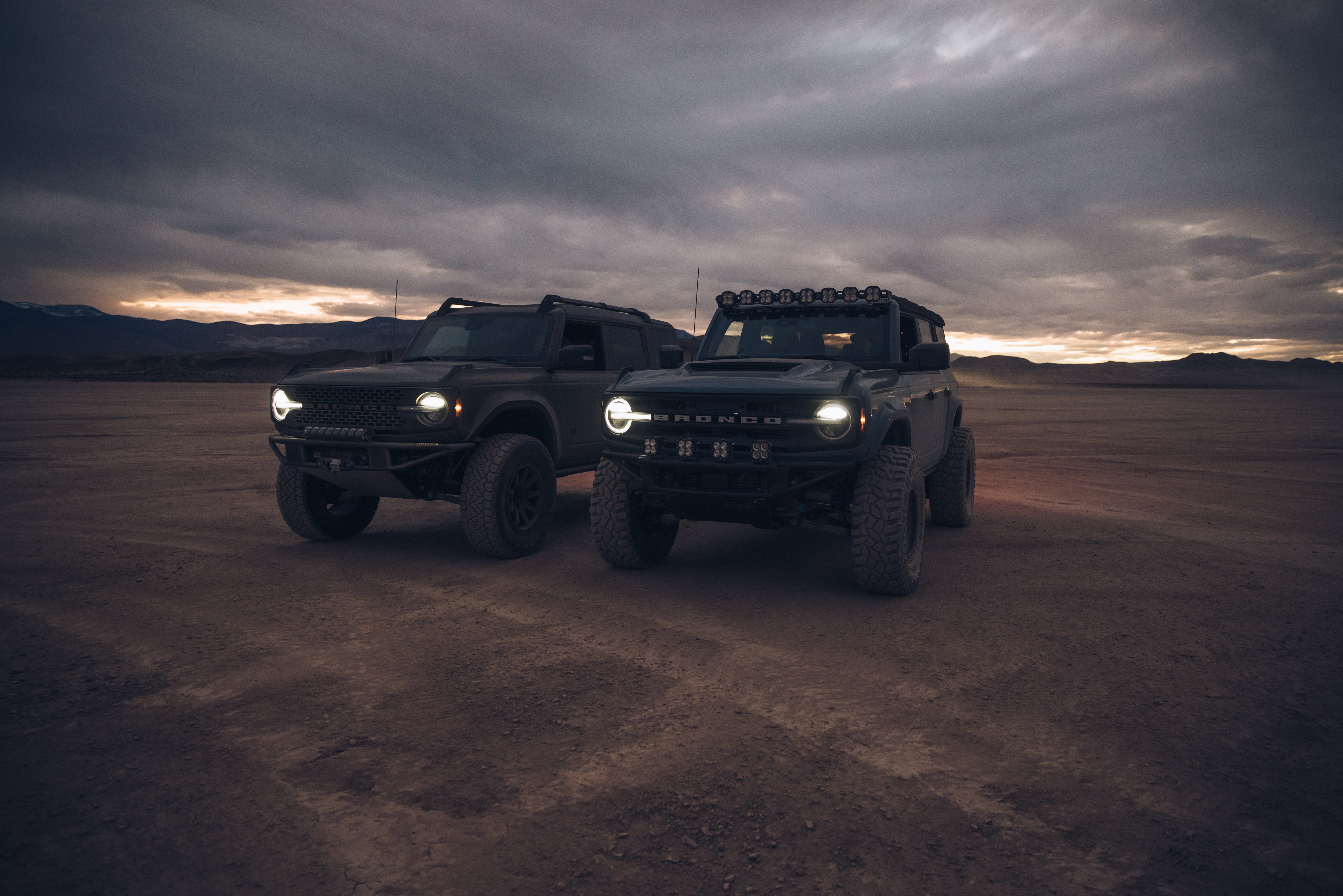 Two modified 2021 Ford Bronco vehicles parked in a rugged landscape at dusk, showcasing offroad capabilities and custom features.