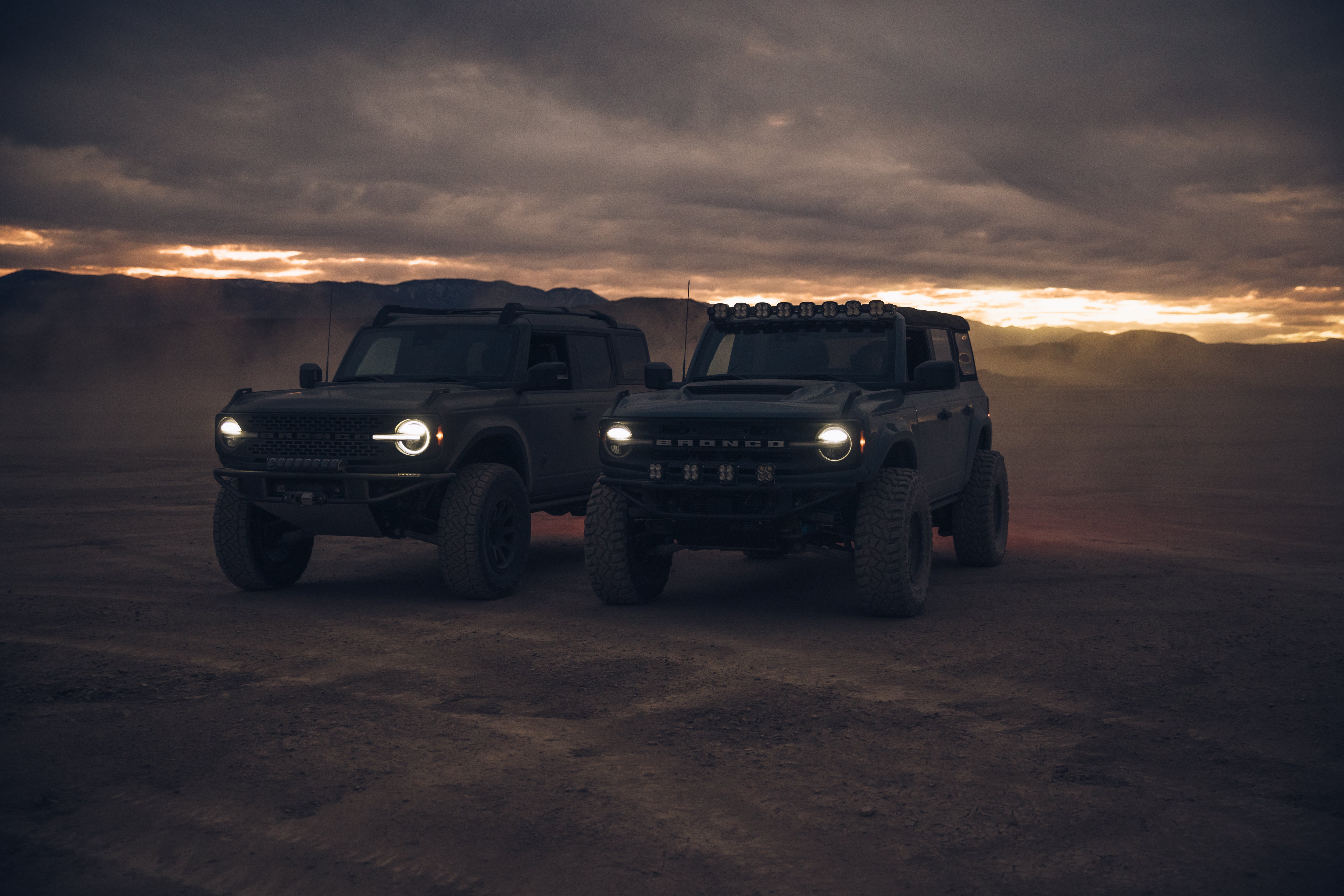 Two 2021 Ford Bronco vehicles parked in a dusty landscape at dusk, showcasing their rugged design and off-road capabilities.