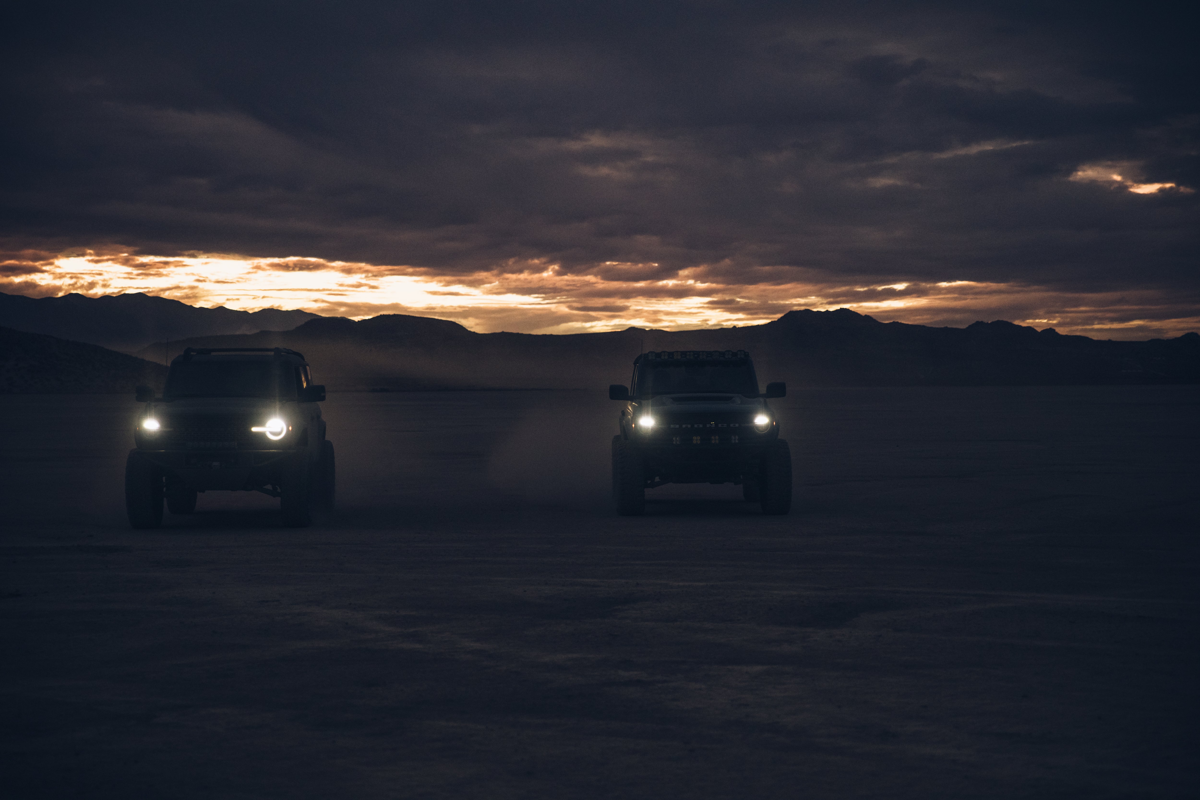 Two 2021 Ford trucks with ADV fiberglass parts driving on a salt flat at sunset, showcasing offroad capabilities.