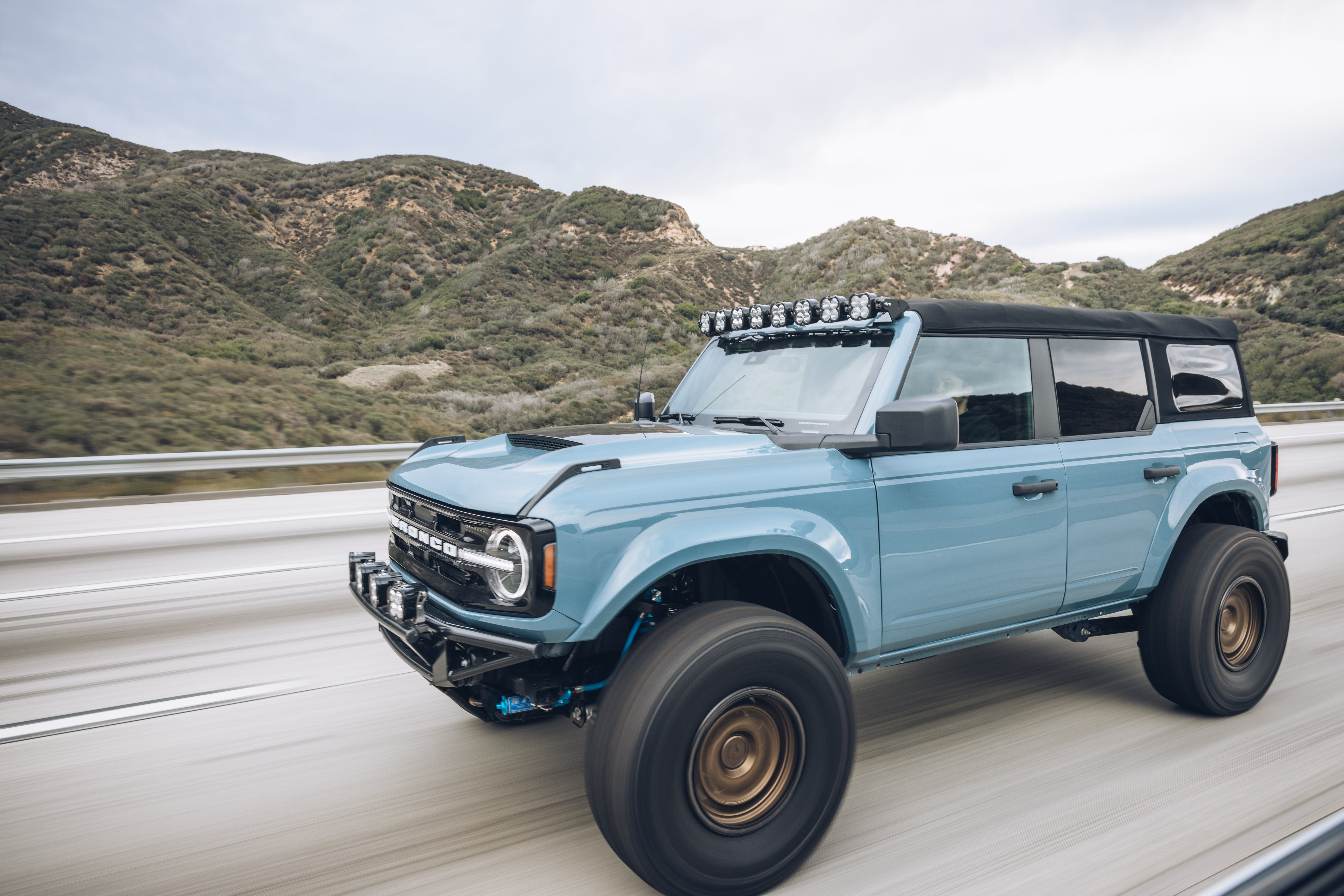 ADV Fiberglass 2021 Ford Bronco with offroad modifications driving on a highway surrounded by hills.