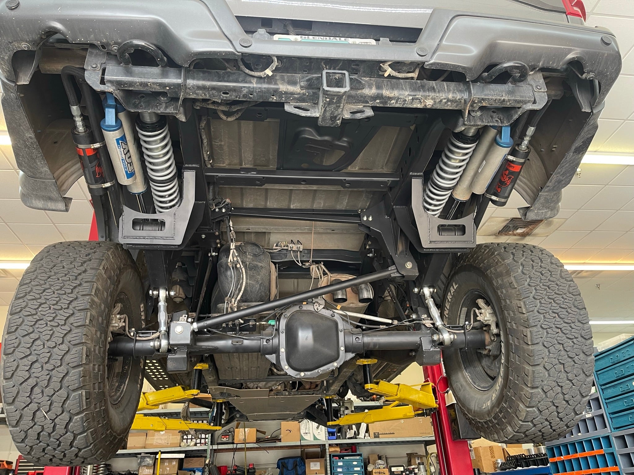 2021-2024 Raptor Long Travel Rear Suspension Kit showcasing 19 inches of travel under a lifted truck in a workshop.