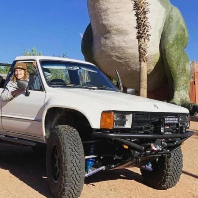 White 1984-1988 Toyota Pickup with aftermarket fenders parked near a large dinosaur statue, showcasing offroad capabilities.