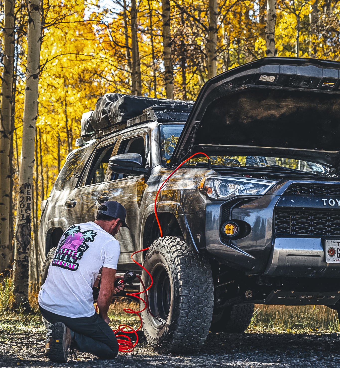 A person inflating a tire on a Toyota 4Runner in a forest setting, wearing a BBO Miami Vice T-Shirt.
