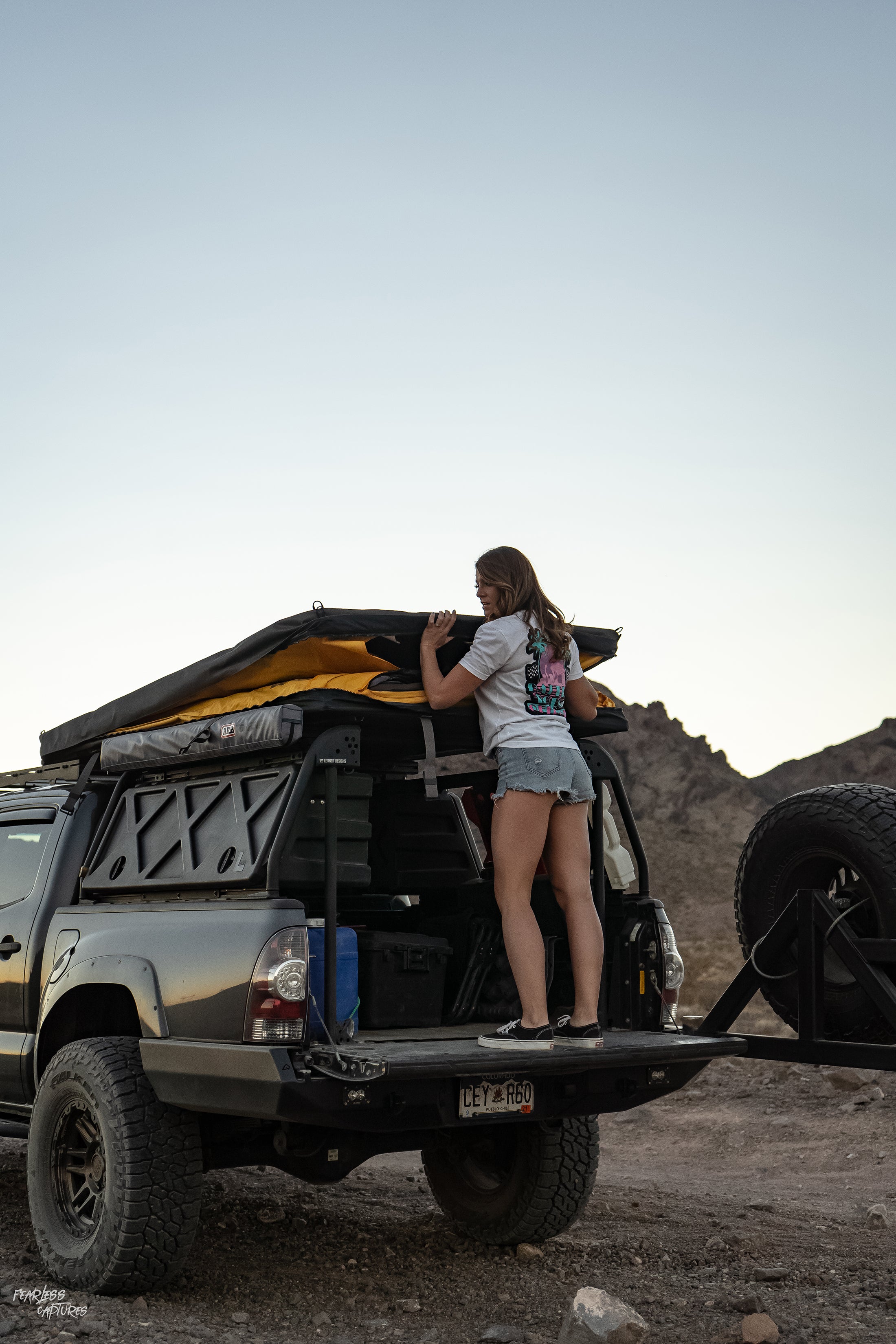 A person wearing a BBO Miami Vice T-Shirt adjusts a rooftop tent on a black SUV in a desert setting.