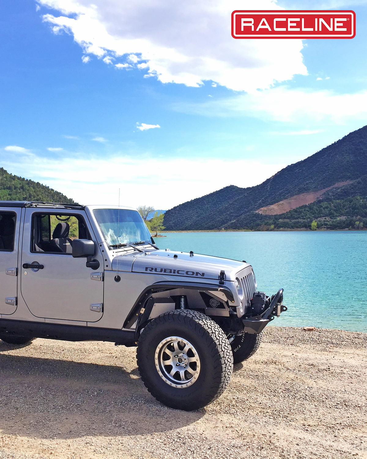 Silver Jeep Rubicon with 929M Avenger wheels parked by a scenic lake, surrounded by mountains and clear blue skies.