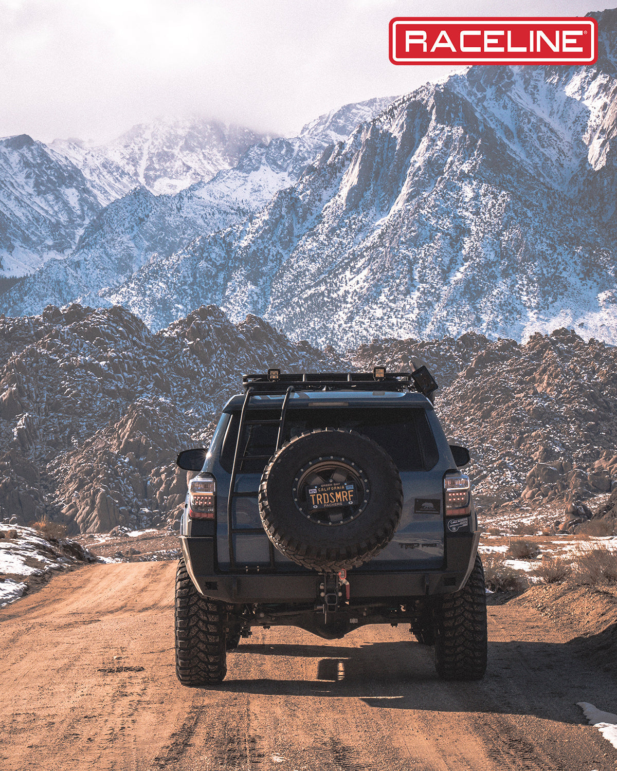 Rear view of a modified SUV on a rugged mountain road, showcasing the 937BZ Combat tire and scenic snowy peaks.