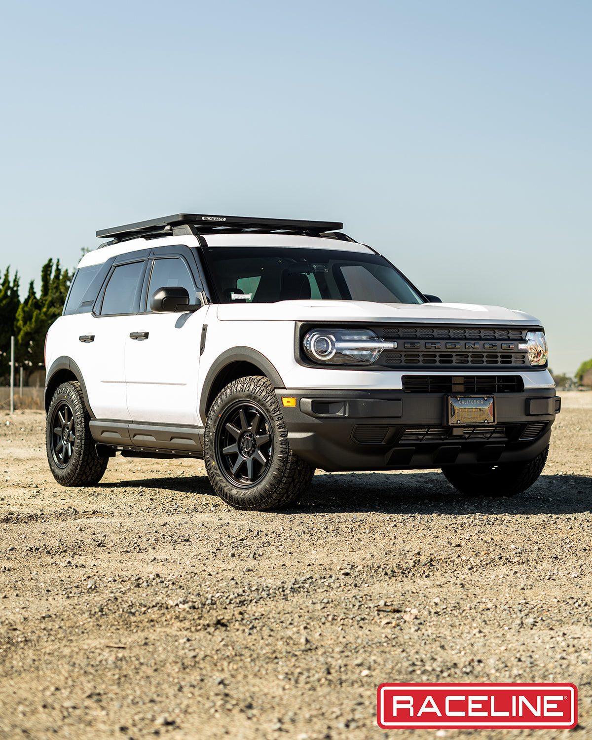 White Ford Bronco with black wheels and roof rack, parked on gravel terrain, showcasing off-road capabilities.