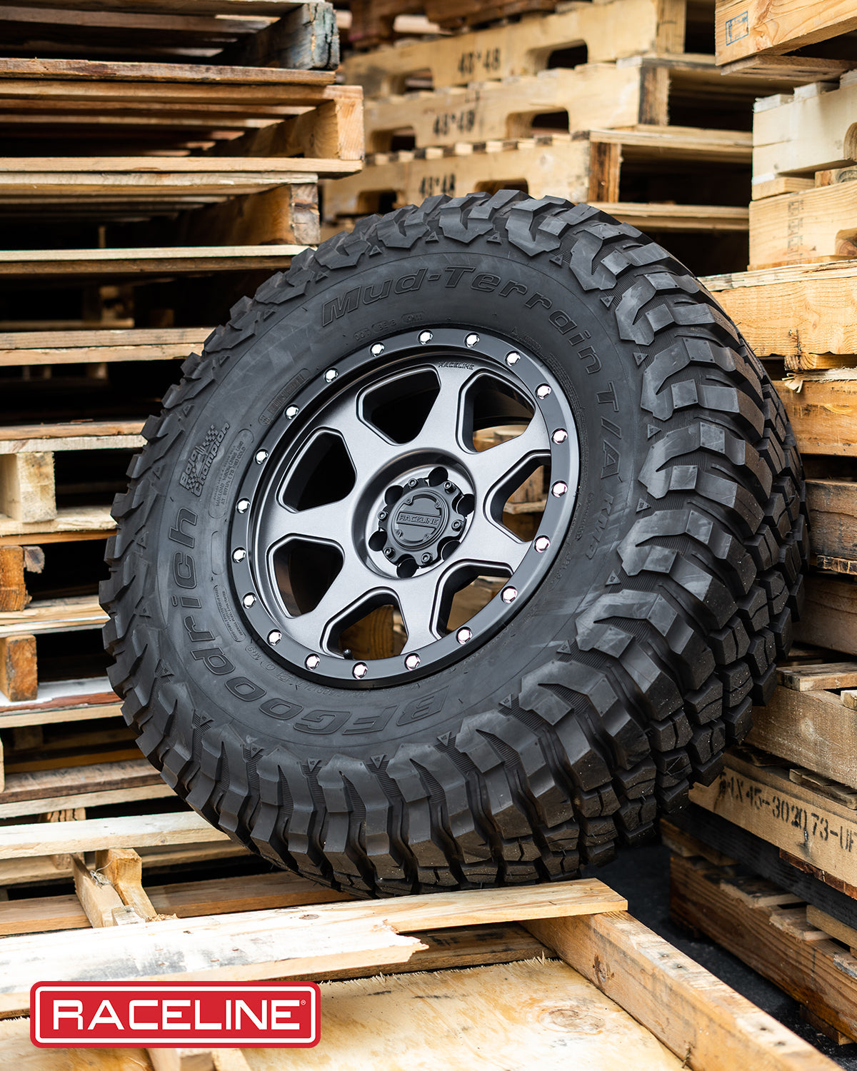 Black off-road tire with a rugged tread pattern mounted on a stylish alloy wheel, displayed among wooden pallets.